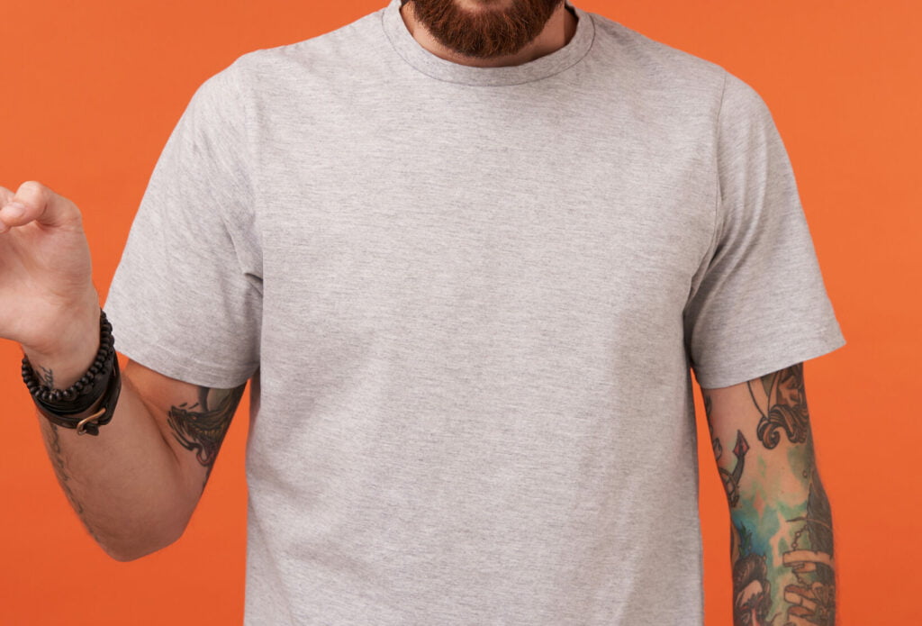Suprised bearded blue-eyed man with tattooes showing aside with index finger and rounding eyes while looking at camera, wearing beige t-shirt while posing over orange background